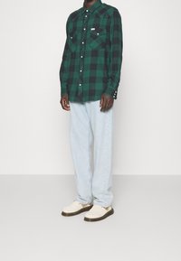 Zielono-czarna koszula w kratę zapinana na guziki, z dwiema kieszeniami na piersi, w połączeniu z jasnoniebieskimi zwężanymi dżinsami i białymi butami bez sznurowadeł.