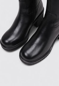 Bottines en cuir noir avec un bout arrondi, une texture lisse, un talon bloc solide et un détail de fermeture éclair sur le côté. Semelle en caoutchouc épais pour une meilleure adhérence.