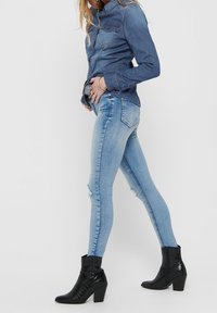 Kvinna iklädd figursydda ljusblå slitna jeans, mörk denimskjorta och svarta klackiga ankelboots, stående på sidan med ena handen i fickan.