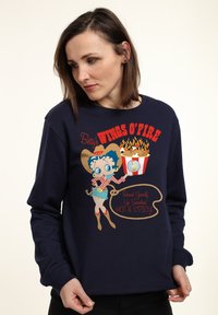 Námořnická mikina s karikaturou ženy v kovbojském oblečení, která drží popcorn. Text zní "Betty's WINGS O'FIRE" s designem plamenů.