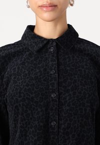 Svart button-up skjorte med et leopardmønster. Har krage, seks knapper og en myk tekstur.