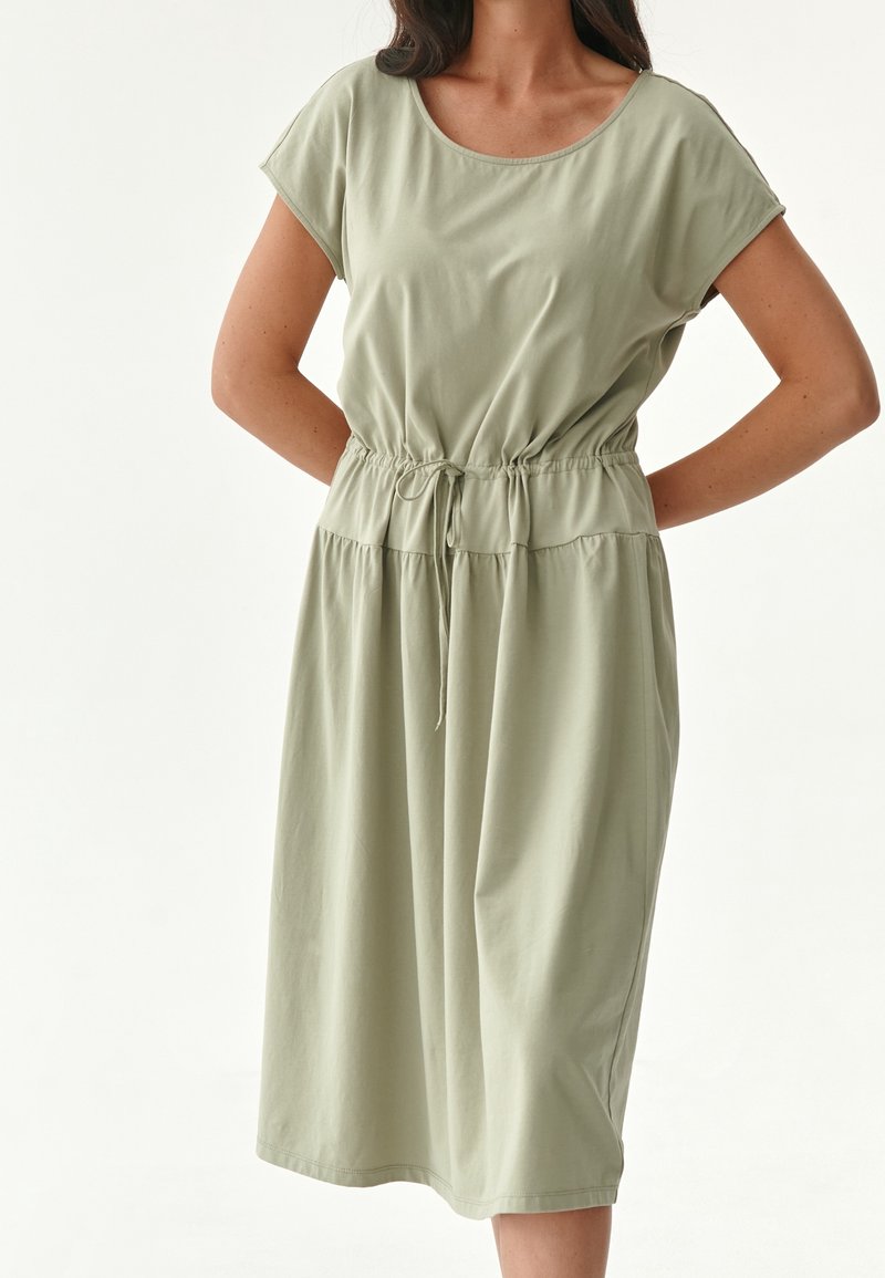 Femme portant une robe verte claire à manches courtes, longueur genou, avec un cordon à la taille, debout les mains derrière le dos.