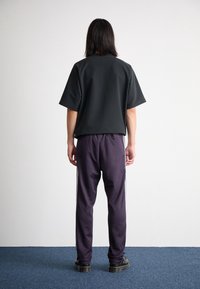 Person med axellångt mörkt hår står vänd mot vit vägg, iförd svart oversized T-shirt, mörka träningsbyxor med vita ränder och svarta skor.