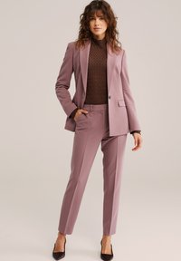 Dopasowany dwuczęściowy garnitur w miękkim odcieniu mauve. Marynarka z wyciętymi klapami i pojedynczym guzikiem; spodnie o wąskim kroju. Pod spodem warstwowy brązowy, fakturowany golf.