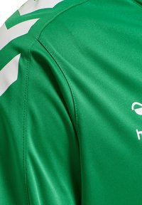 Close-up van een groen sportshirt met zichtbare schoudernaad, stoftextuur, wit chevrondesign en gedeeltelijk wit logo op de borst.