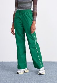 Πράσινα νάιλον παντελόνια με χαλαρή εφαρμογή, ελαστική μέση και λευκές λεπτομέρειες στα πλάγια. Το μοντέλο φορά ασημένια αθλητικά παπούτσια με γκρι λεπτομέρειες.