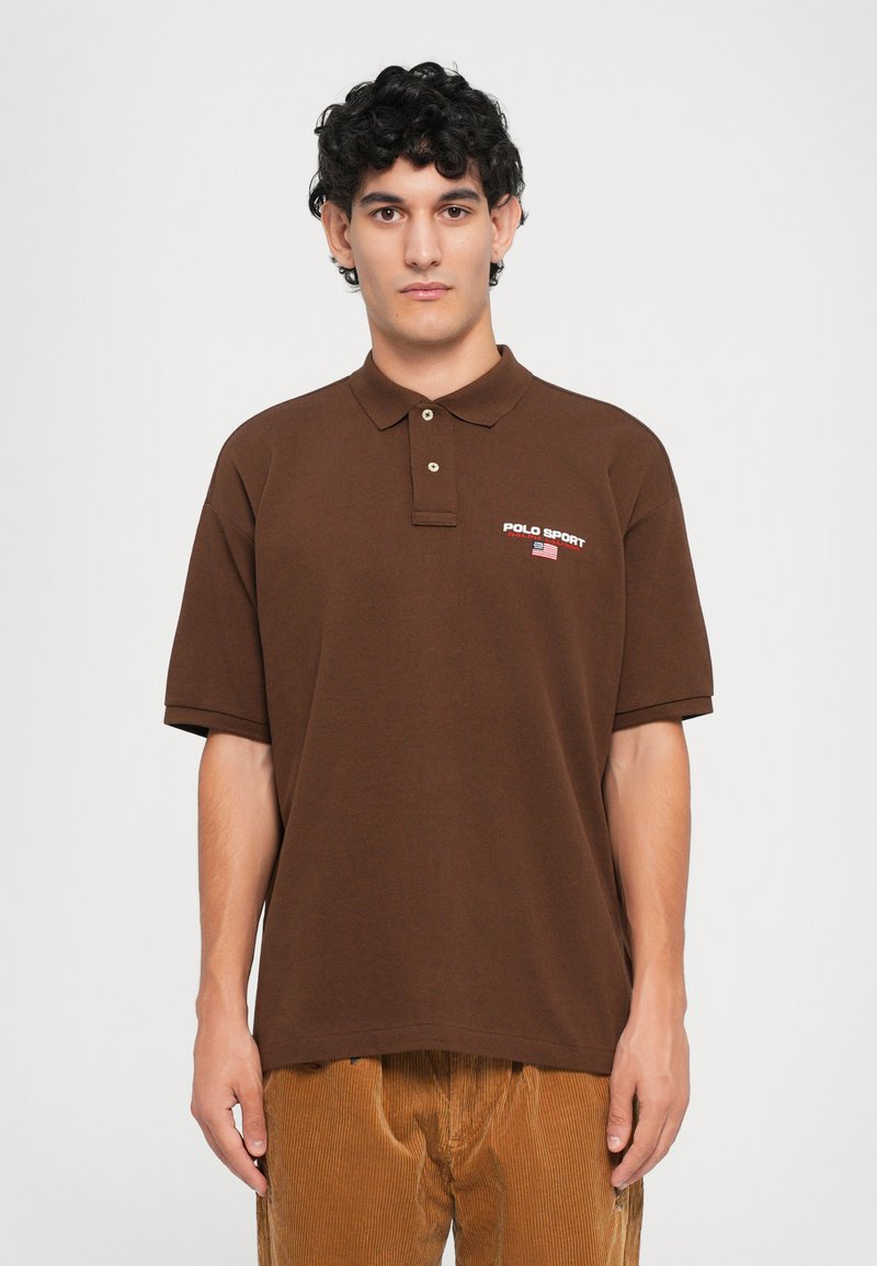 Polo castanho feito de algodão, com colarinho e dois botões, apresentando a marca "POLO SPORT" em vermelho no lado esquerdo do peito. Design simples.