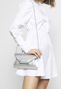Bolso de hombro metálico plateado con un solapa triangular, correa de cadena y una textura suave. Sostenido por una persona que lleva un vestido blanco.