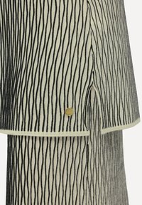 Indumento lavorato a maglia con strisce nere verticali su uno sfondo crema. Comprende un dettaglio con distintivo dorato e uno spacco corto all'orlo.