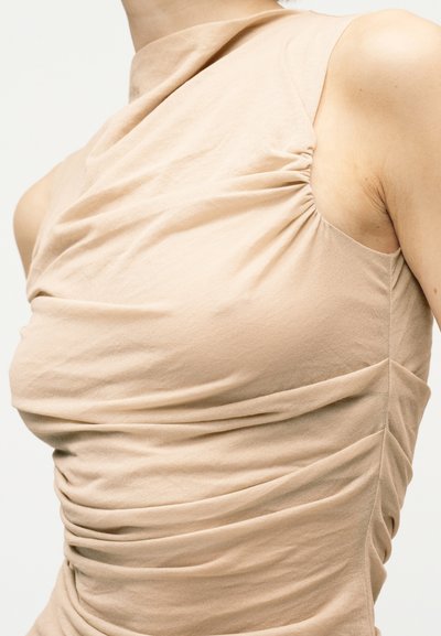 Μπεζ αμάνικη μπλούζα με ψηλό λαιμόκοψη και ρυτιδώσεις στο στήθος και τις πλευρές, φορεμένη από ένα άτομο σε απλό φόντο.