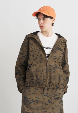 Junge Person trägt eine orangefarbene Kappe, ein weißes T-Shirt mit Grafik, eine braun gemusterte Reißverschlussjacke und passende Hose, die Hände in den Taschen, schaut nach rechts.
