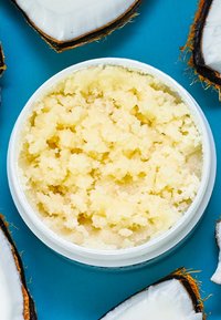 Scrub al cocco in un contenitore rotondo bianco. Lo scrub testurizzato è di un giallo pallido, circondato da gusci di cocco su uno sfondo blu.