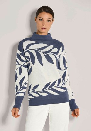 Vrouw in een blauw-wit trui met grote bladerenpatronen en een witte broek, staande tegen een effen lichtgrijze achtergrond.