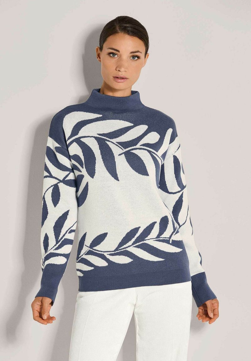 Frau in einem blau-weißen Pullover mit großen Blattmustern und weißen Hosen, die vor einem einfarbigen hellgrauen Hintergrund steht.