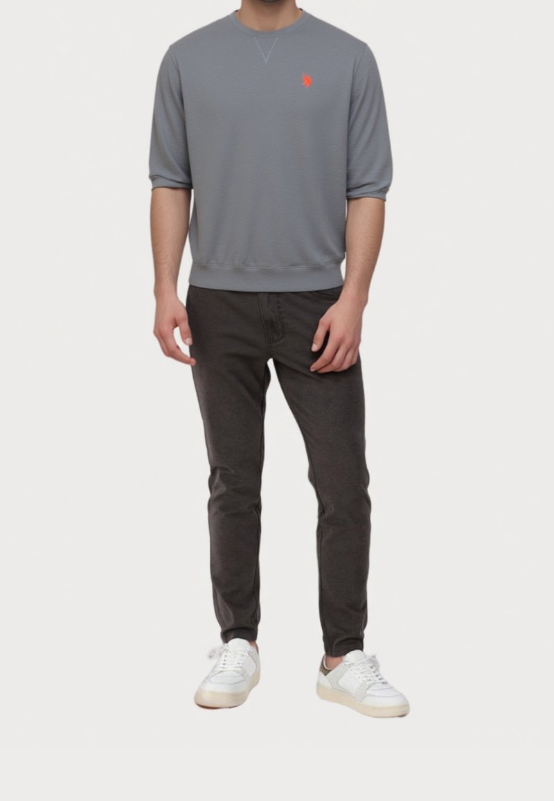 Homme portant un sweat-shirt gris à manches courtes avec un logo orange, un pantalon noir et des baskets blanches, debout devant un fond uni.
