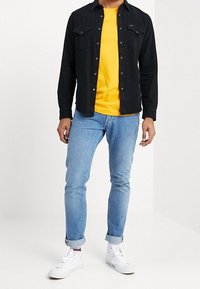 Musta pitkähihainen paita keltaisen t-paidan päällä, yhdistettynä vaaleansinisiin skinny-farkkuihin ja valkoisiin lenkkereihin punaisilla yksityiskohdilla.