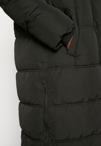 Černý prošívaný kabát s výrazným prošíváním, vybavený bočními kapsami, elastickými manžetami a zipem na spodním lemu.