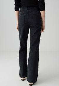 Persoon in hoge taille zwarte rechte jeans en zwarte lange mouwen top, staand op een lichtgrijze vloer tegen een effen achtergrond.