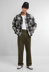 Zwart-wit geruite flanellen shirt met twee voorzakken, gecombineerd met olijfgroene wijde broek en een zwarte gebreide beanie. Witte sneakers zijn ook zichtbaar.