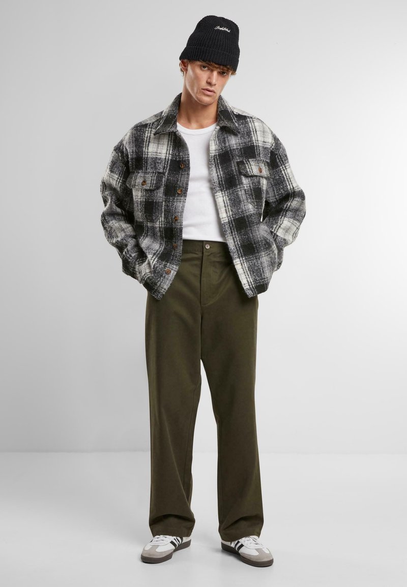 Zwart-wit geruite flanellen shirt met twee voorzakken, gecombineerd met olijfgroene wijde broek en een zwarte gebreide beanie. Witte sneakers zijn ook zichtbaar.