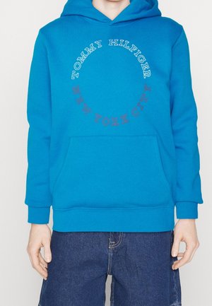Osoba nosząca jasnoniebieską bluzę z kapturem Tommy Hilfiger z napisem "Tommy Hilfiger New York City" w okrągłym wzorze z przodu.