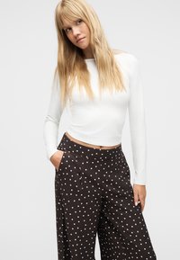 Νεαρή γυναίκα με μακριά ξανθά μαλλιά που φοράει κολλητό λευκό μακρυμάνικο τοπ και μαύρο παντελόνι με φαρδιά πόδια και λευκές πουά, με τα χέρια στις τσέπες.