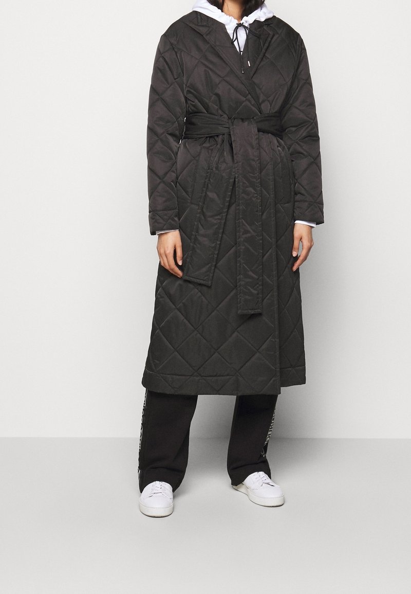 Personne portant un manteau noir matelassé ceinturé par-dessus un sweat à capuche blanc, un pantalon noir et des baskets blanches, debout devant un fond uni.