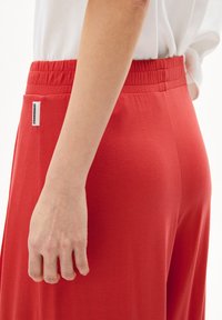 Rode, elastische taillebandbroeken met een gladde textuur. Heeft een zijlabel met branding. Afbeelding toont de tailleband en de handplaatsing.