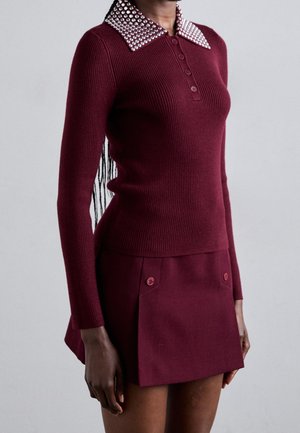 Kvinde iført en tætsiddende bordeaux rød striktrøje med polkaprikket krave og matchende mininederdel med knappede forlommer.