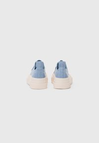 Blue denim slip-on shoes with a creamy white rubber sole, featuring a textured finish and small fabric loops on the heel.