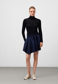 Marinblå veckad kjol med bred midjeband och knytbälte, i kombination med en figurnära svart polotröja och beige sneakers på ett grått golv.