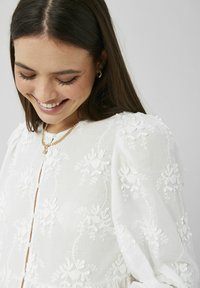 Blouse en lin blanc avec broderies florales, col rond et fermeture par boutons. Comprend des manches ballon et des motifs texturés délicats.