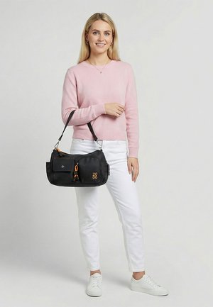 Kobieta w różowym swetrze i białych spodniach trzyma czarną torbę na ramię z pomarańczowymi akcentami i numerem "2006".