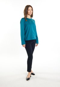 Türkiser Langarm-Oberteil mit Verzierungen an den Schultern, kombiniert mit schwarzen Slim-Fit-Hosen und spitzen schwarzen Ballerinas, vor einem weißen Hintergrund.
