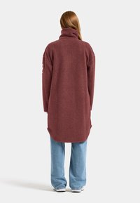 Bordeaux, zachte fleece jas met een hoge kraag, lange mouwen en een langere achterkant, gecombineerd met lichtblauwe wijde jeans.