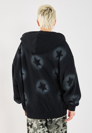 Osoba z krótkimi blond włosami, ubrana w czarną bluzę z kapturem z wyblakłymi wzorami gwiazd i spodnie moro, zwrócona tyłem na jednolitym tle.