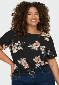 Top floreale nero con maniche corte, caratterizzato da fiori rosa e bianchi, indossato con jeans blu e una cintura nera con fibbia circolare dorata.