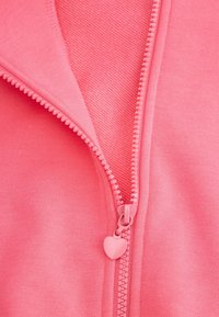 Różowa kurtka zapinana na zamek z gładką fakturą materiału, z zamkiem w kształcie serca oraz kontrastową wewnętrzną warstwą widocznym szwem.