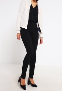Black stretchy skinny jeans paired with a loose black top and a light bomber jacket. Model wearing black patent heels, showcasing a sleek silhouette.