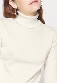 Cream turtleneck sweater made of soft knit fabric, featuring a snug collar and ribbed texture. Simple design with long sleeves.