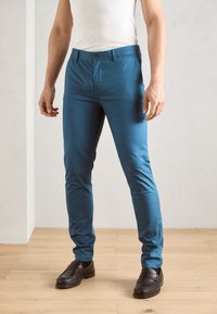 Moški v slim-fit modrih hlačah, črnih loaferjih in belem brezrokavniku stoji na lesenem tlu znotraj.