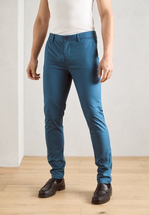 Mann in schlanken blauen Hosen, schwarzen Slippern und einem weißen ärmellosen Hemd, der auf einem Holzfußboden drinnen steht.