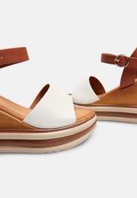 Sandali platform in pelle con una larga fascia bianca, cinturino marrone alla caviglia, suola intermedia in legno e suola in schiuma stratificata per un'altezza e comfort aggiuntivi.
