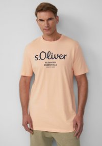 Licht perzikkleurig katoenen T-shirt met een zwarte opdruk van de merknaam "s.Oliver" en "Elevated Essentials" gecentreerd op de voorkant. Korte mouwen.