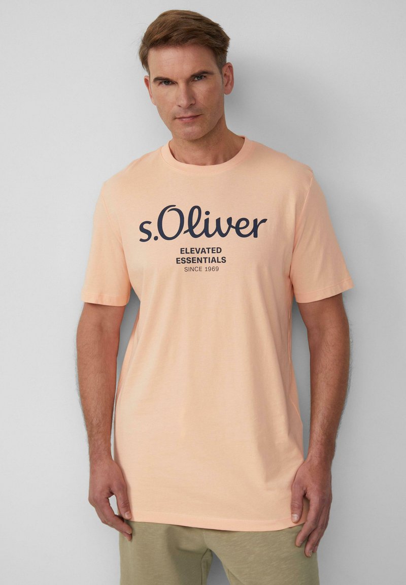 Licht perzikkleurig katoenen T-shirt met een zwarte opdruk van de merknaam "s.Oliver" en "Elevated Essentials" gecentreerd op de voorkant. Korte mouwen.