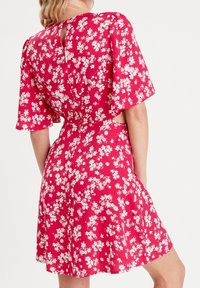Robe florale rouge à manches courtes, avec des découpes sur les côtés et un dos en forme de trou de serrure. Présente des motifs de fleurs blanches et une jupe évasée.