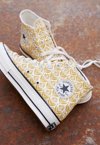 Bílé vysoké tenisky s žlutými listovými vzory, s černým hvězdicovým logem, bílými tkaničkami a gumovou podrážkou s tmavými akcenty.