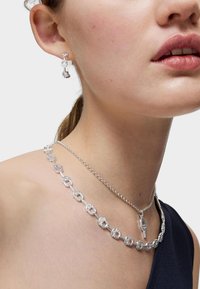 Joyería de plata que presenta un collar de múltiples eslabones con un charm de candado y pendientes a juego con acentos de cristal, complementando un atuendo oscuro.