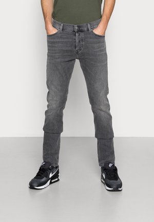 Mężczyzna w wyblakłych szarych dżinsach z rękami w kieszeniach i czarnych butach Nike Air Max stoi na szarej podłodze na tle białej ściany.