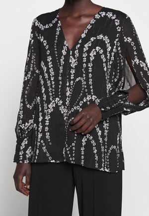 Blouse noire avec motifs floraux blancs en spirale, col en V et manches longues avec fentes, portée avec un pantalon noir sur une personne à la peau foncée.